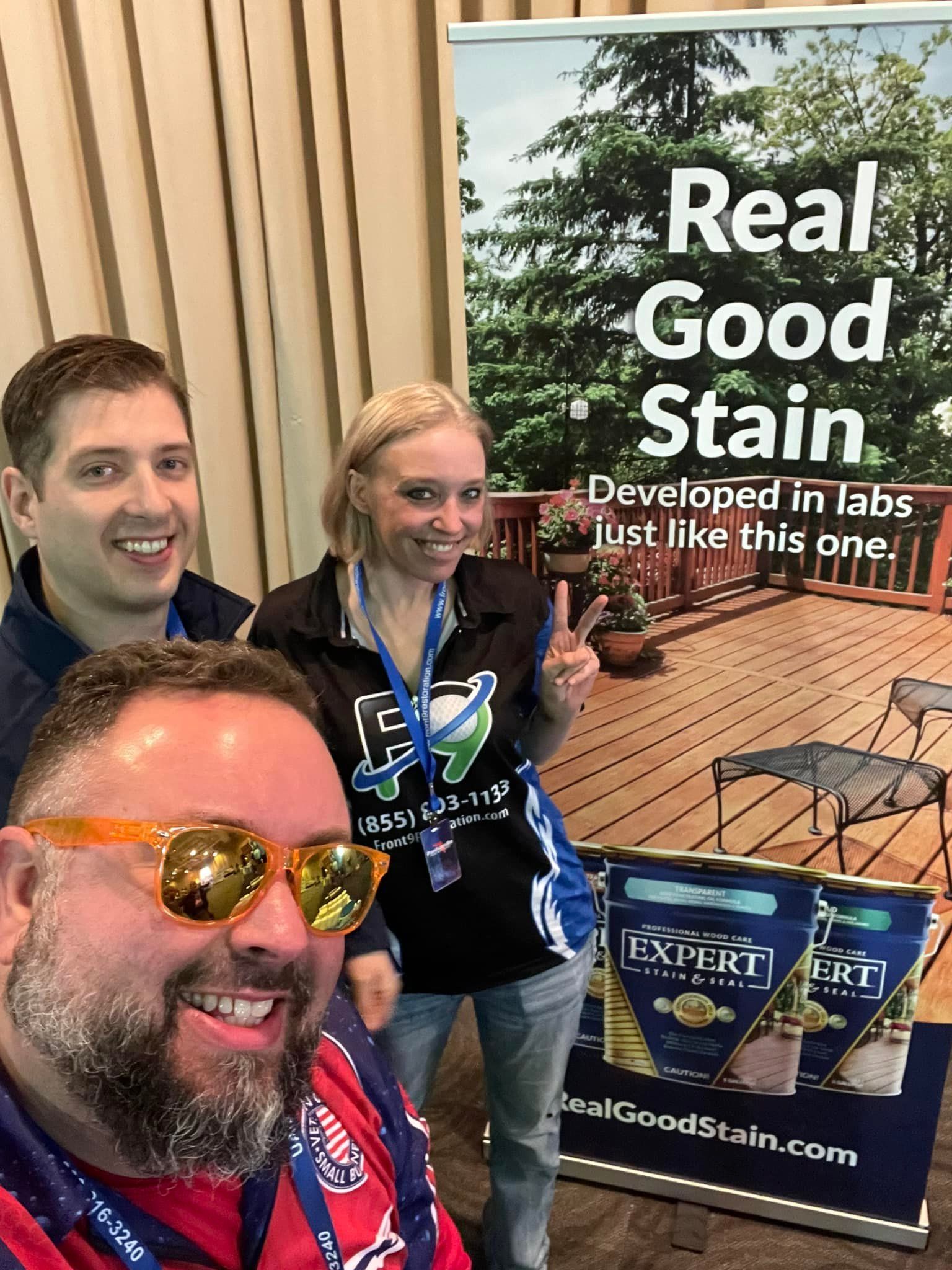 Three people are posing for a picture in front of a sign that says `` real good stain ''.
