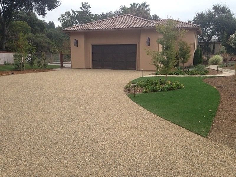 A beige gravel driveway leads to a tan garage with a brown door; green grass borders the drive.