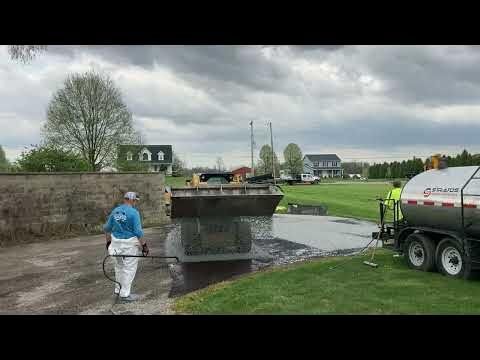 Workers sealcoating a driveway, one spraying, the other by truck, homes in background.