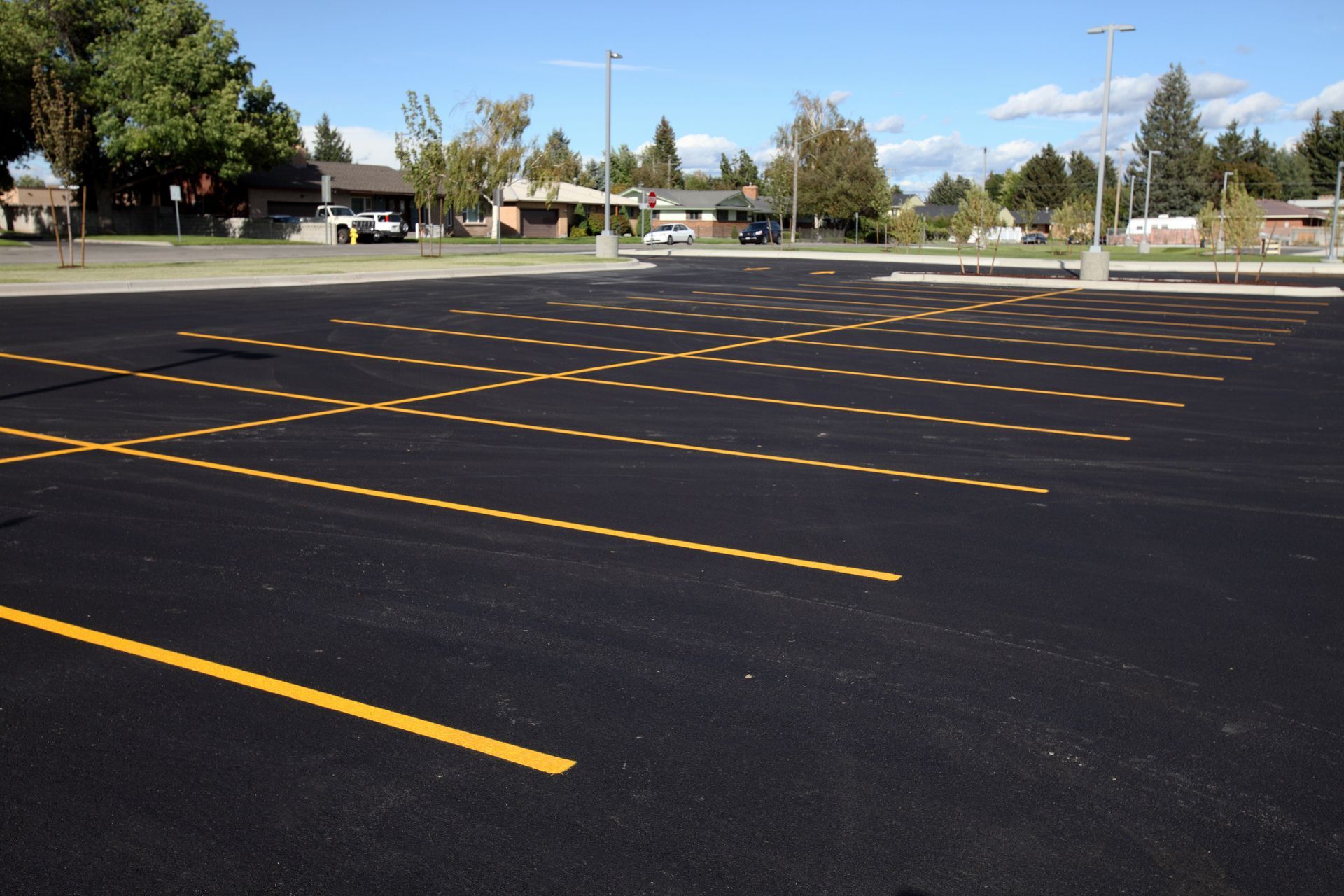 Empty asphalt parking lot with yellow parking space lines, with houses and trees in the background.