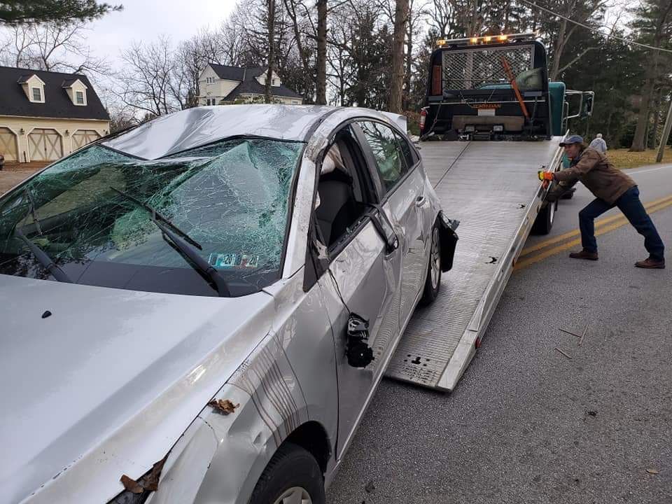 A silver car with a broken windshield is being towed by a tow truck