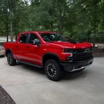 Red pickup truck parked on a driveway beside trees and grass.