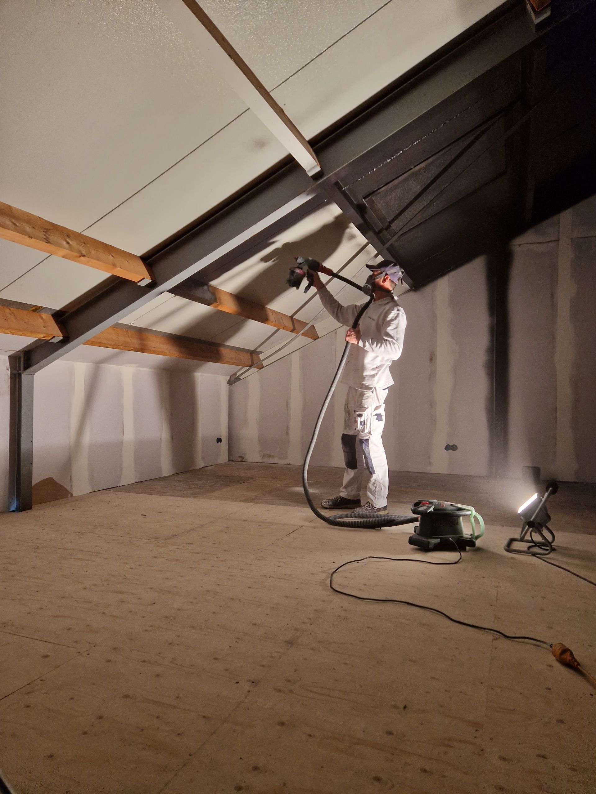 Stefan Go3 spuit verf op een schuin plafond in een kamer met houten balken en gipsplaat.