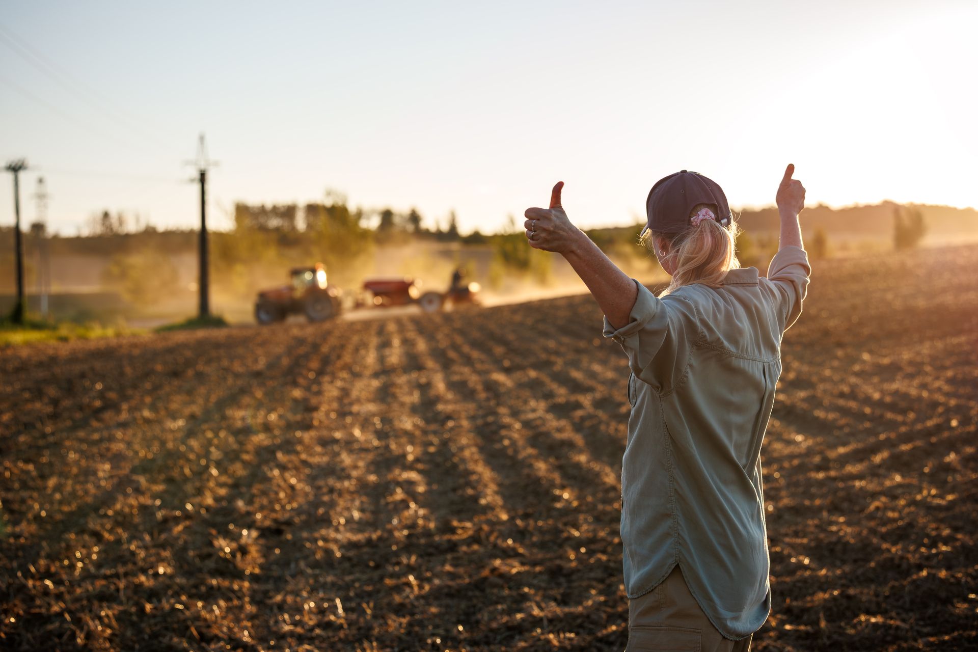 A farmer raising her arms with thumbs up after a sowing process by agricultural machinery. A farmer raising her arms with thumbs up after a sowing process by agricultural machinery.