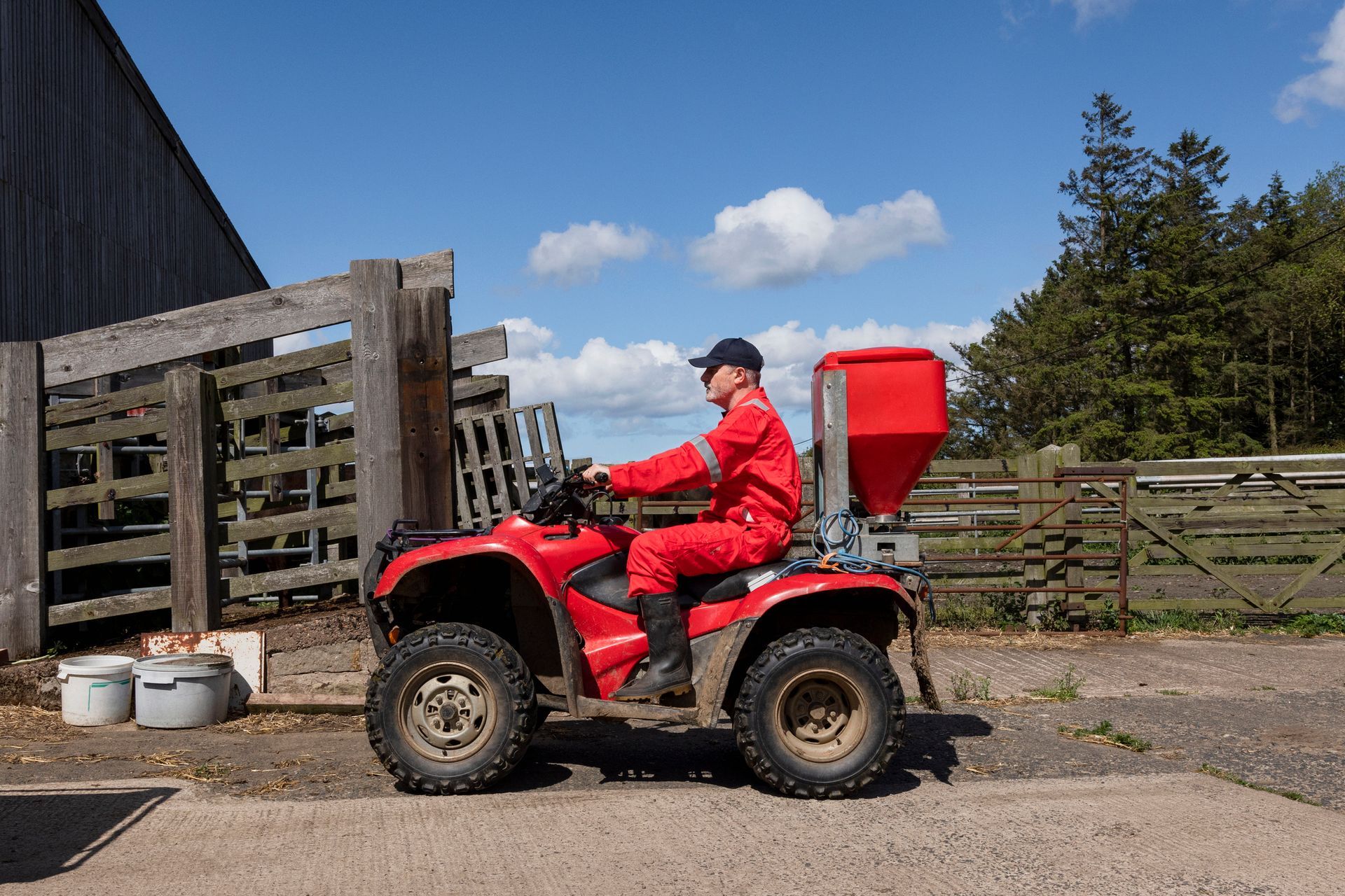 A farmer wearing red overalls is sitting on an ATV vehicle in a farmyard.