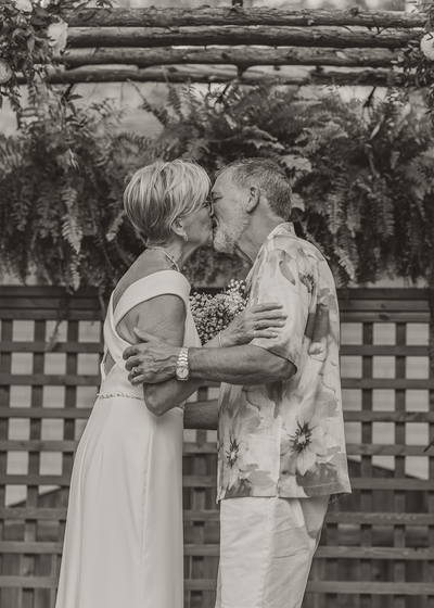 Two women kissing outdoors in front of a rustic wooden wall and greenery