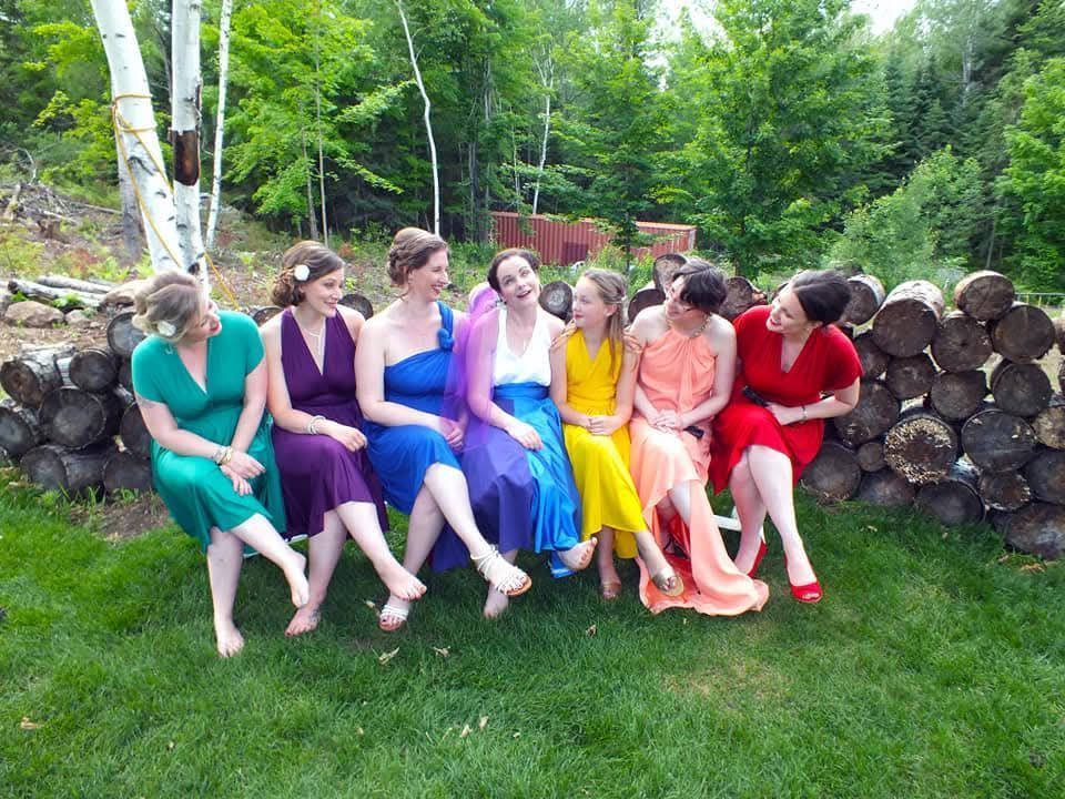 Seven people in colorful dresses sit on a bench outdoors beside stacked logs and trees