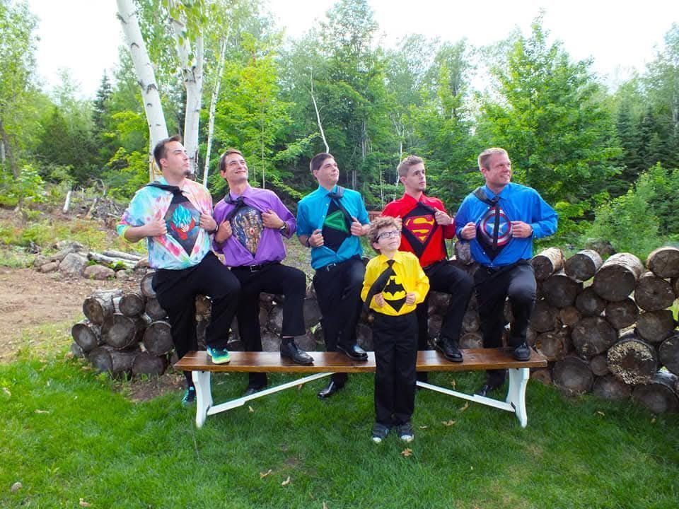 Five people in colorful shirts sit on a wooden bench outdoors, smiling and playing instruments near a log pile.