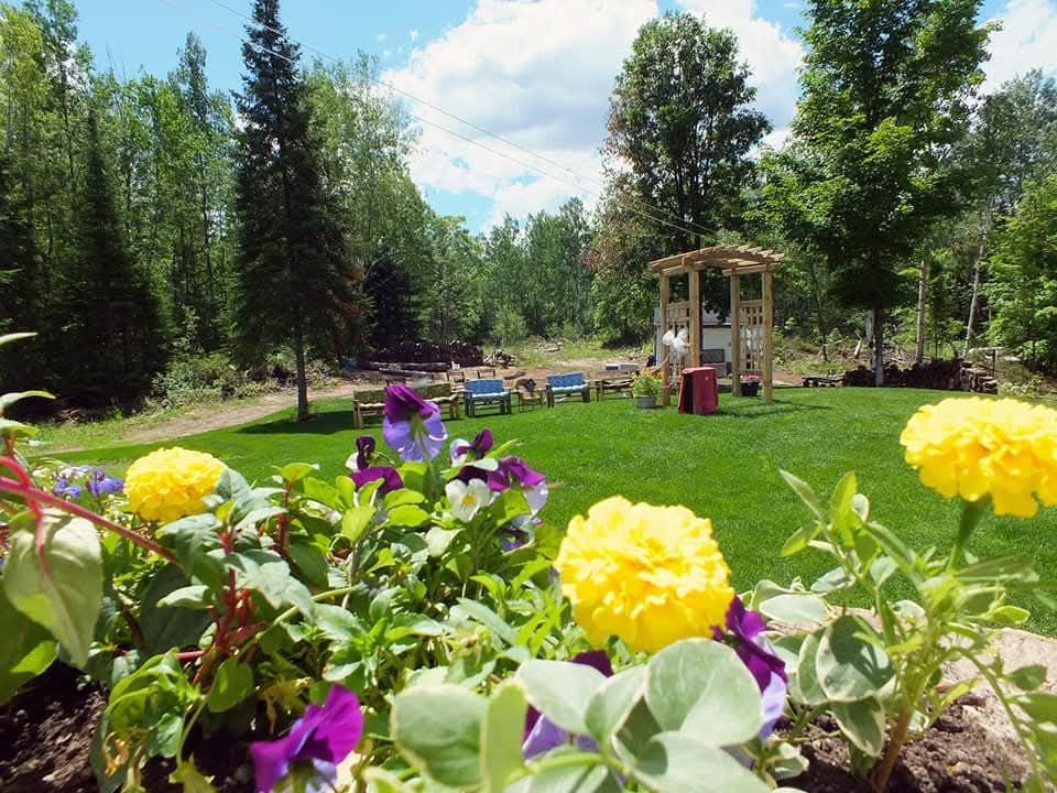 Bright flower garden with yellow and purple blooms, green lawn, trees, and a small gazebo under a blue sky