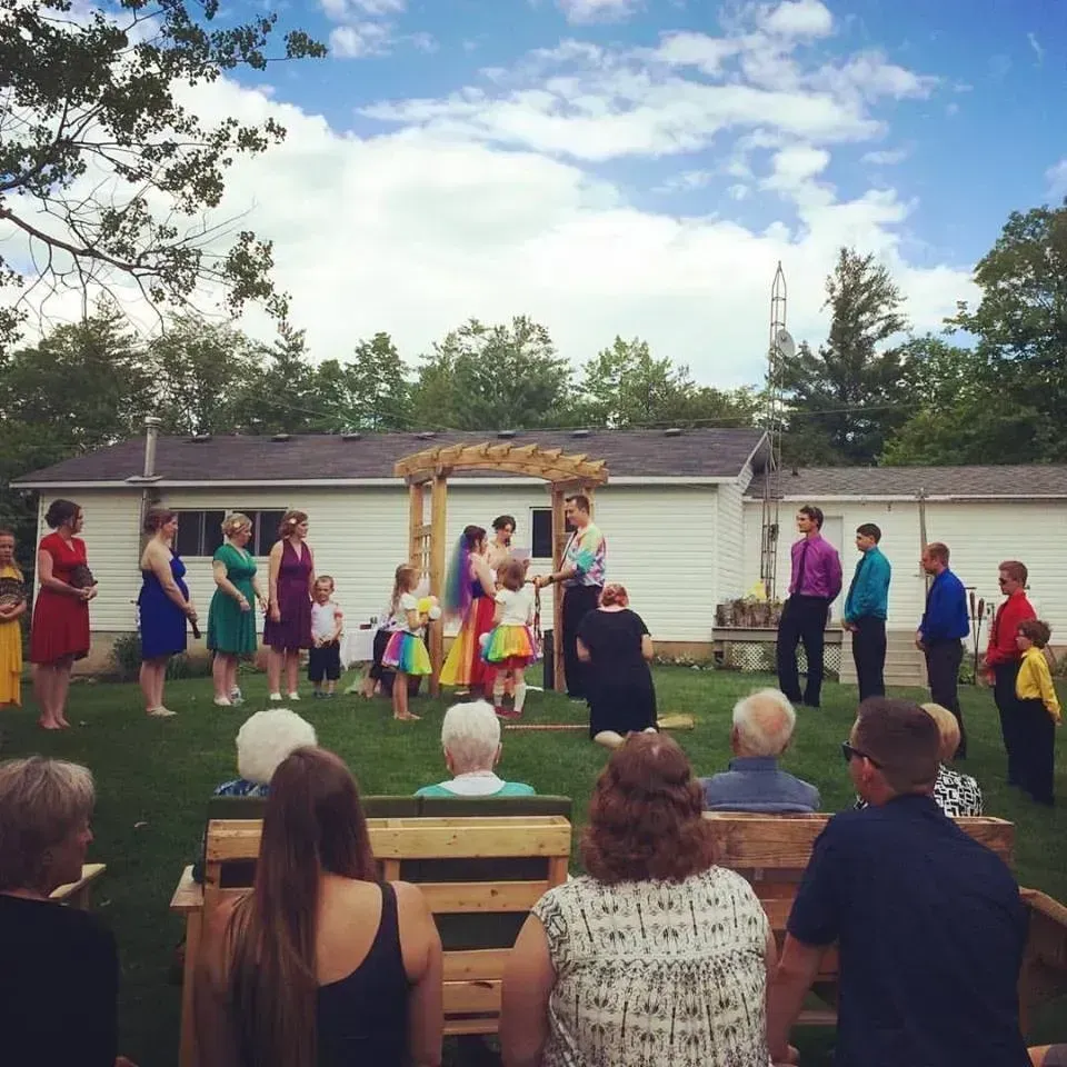 Outdoor performance of people in colorful costumes on a lawn before an audience, with a house and trees behind them