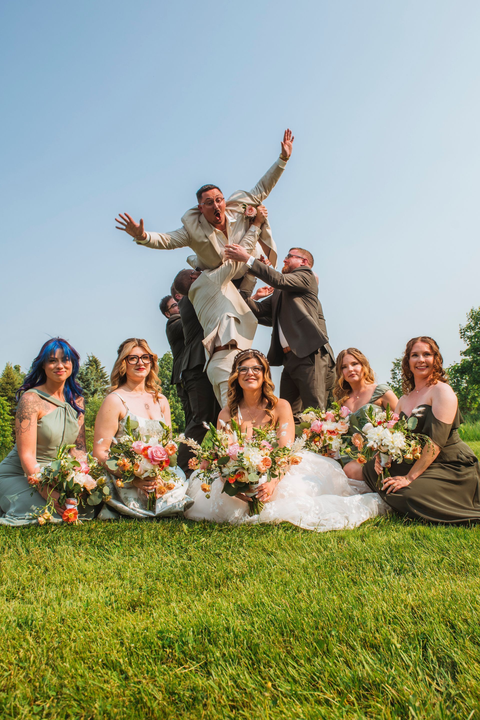Wedding party posing on a grassy lawn, with the couple lifted at center amid bouquets and bright blue sky.