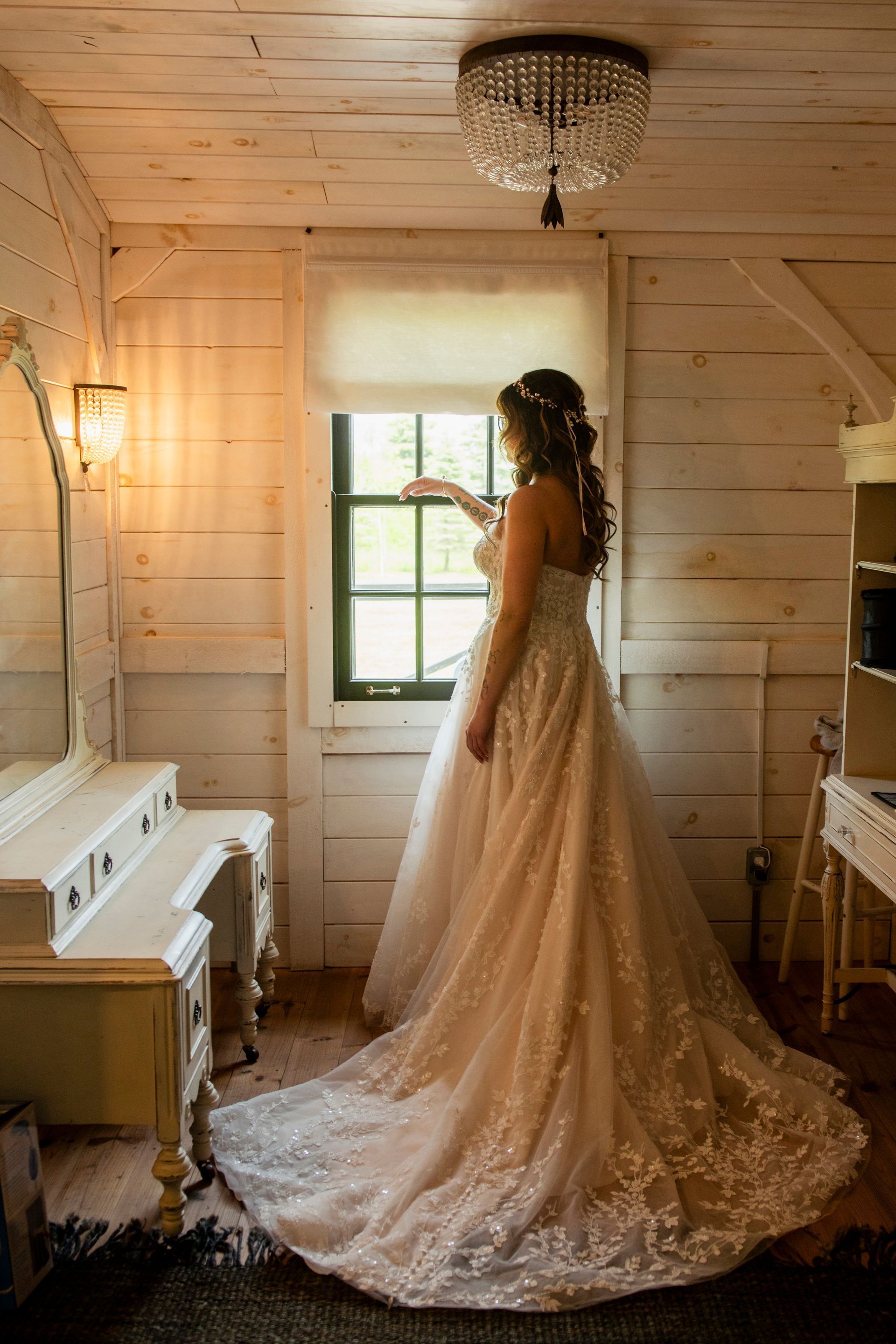 Bride in a lace gown standing by a window in a rustic white dressing room