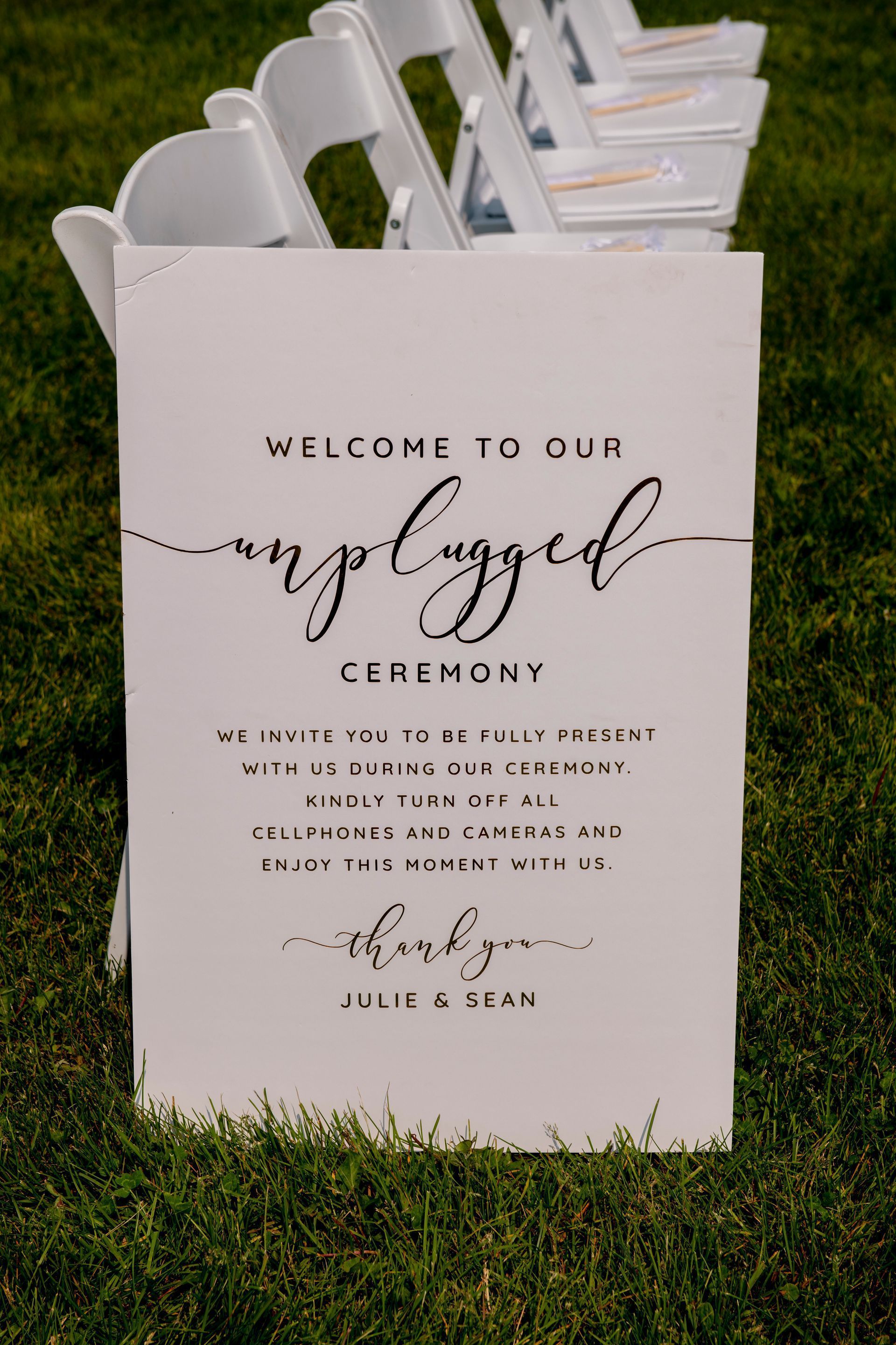 White “Welcome to our unplugged ceremony” sign on grass with stacked white chairs behind it.