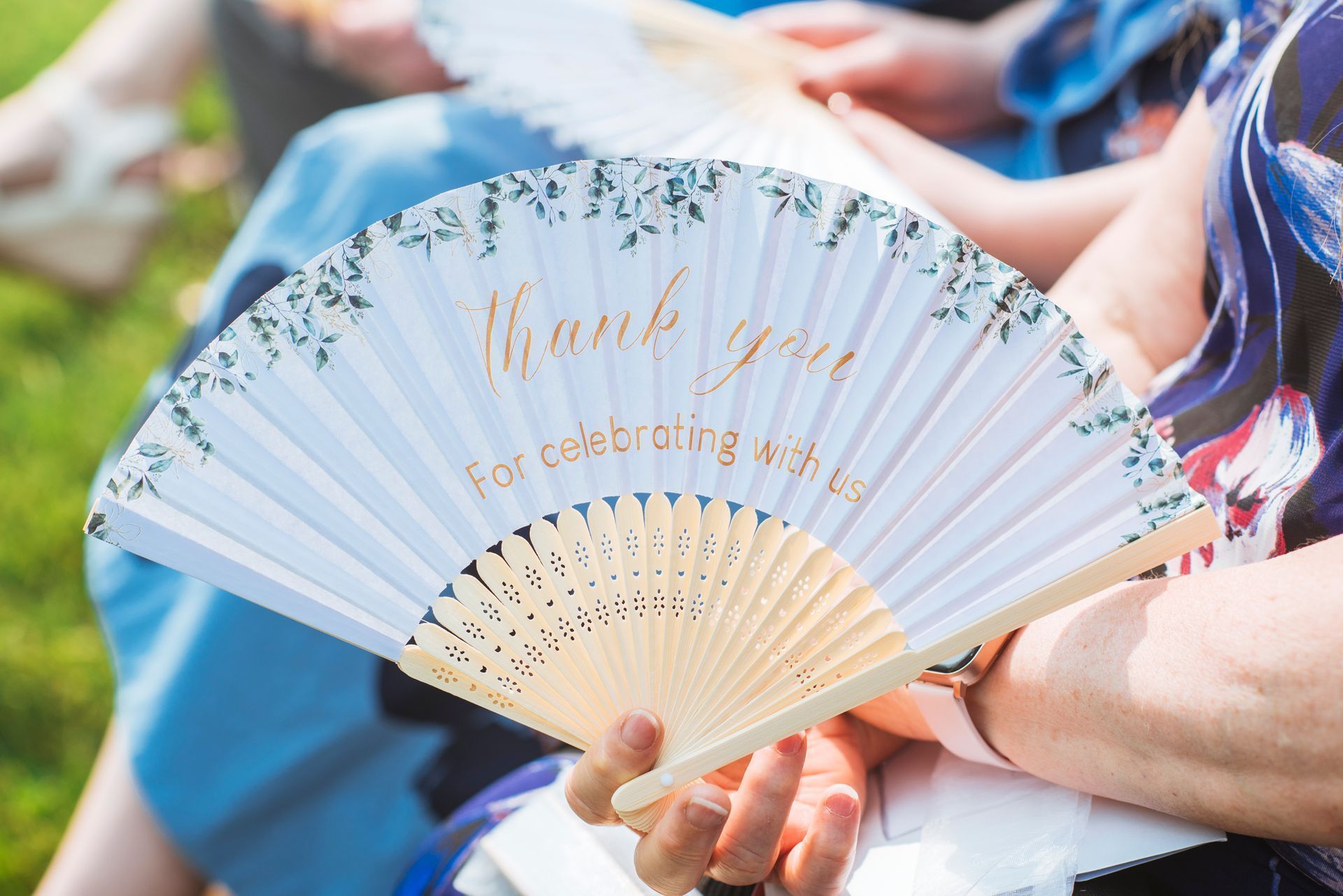White folding fan with gold script held by someone outdoors, with people sitting in the background
