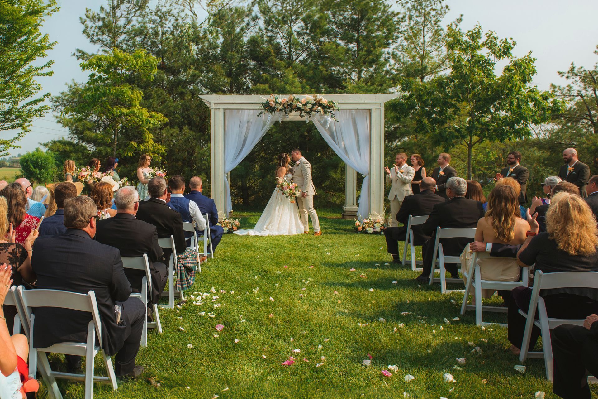 Outdoor wedding ceremony with bride and groom under a white arch, guests seated on both sides of a grass aisle.