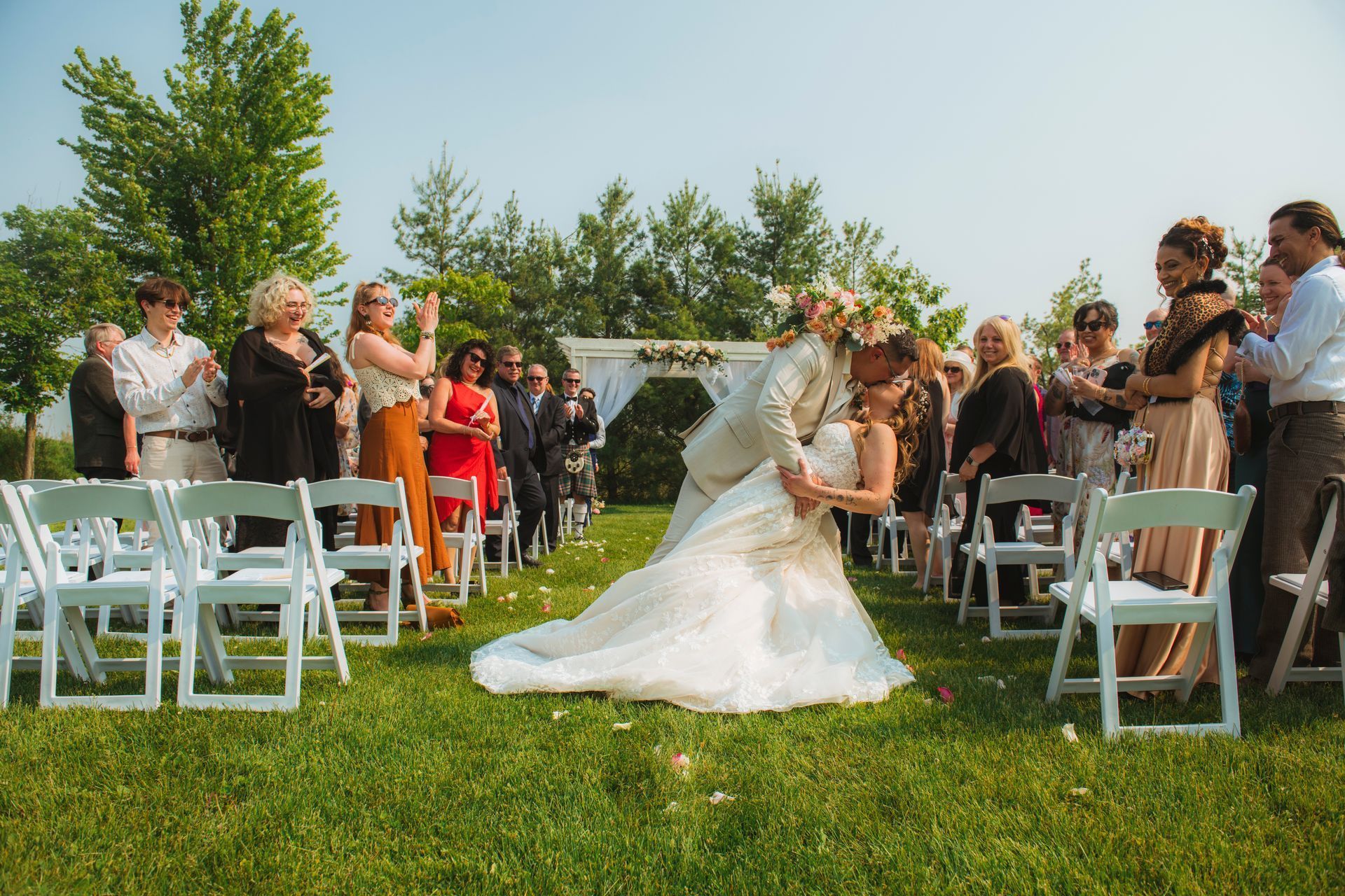 Bride in white gown and groom on grass aisle at outdoor wedding, guests seated on both sides.