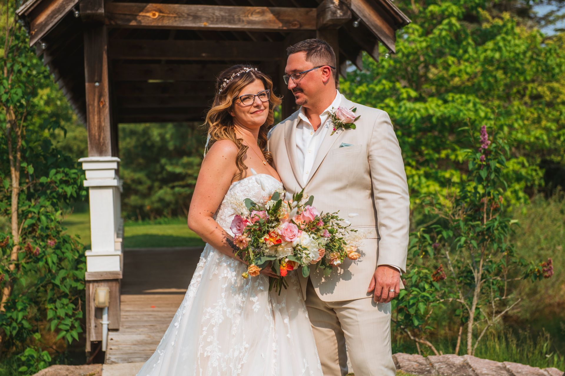 Wedding couple posing under a wooden gazebo, smiling and holding a pastel bouquet outdoors.