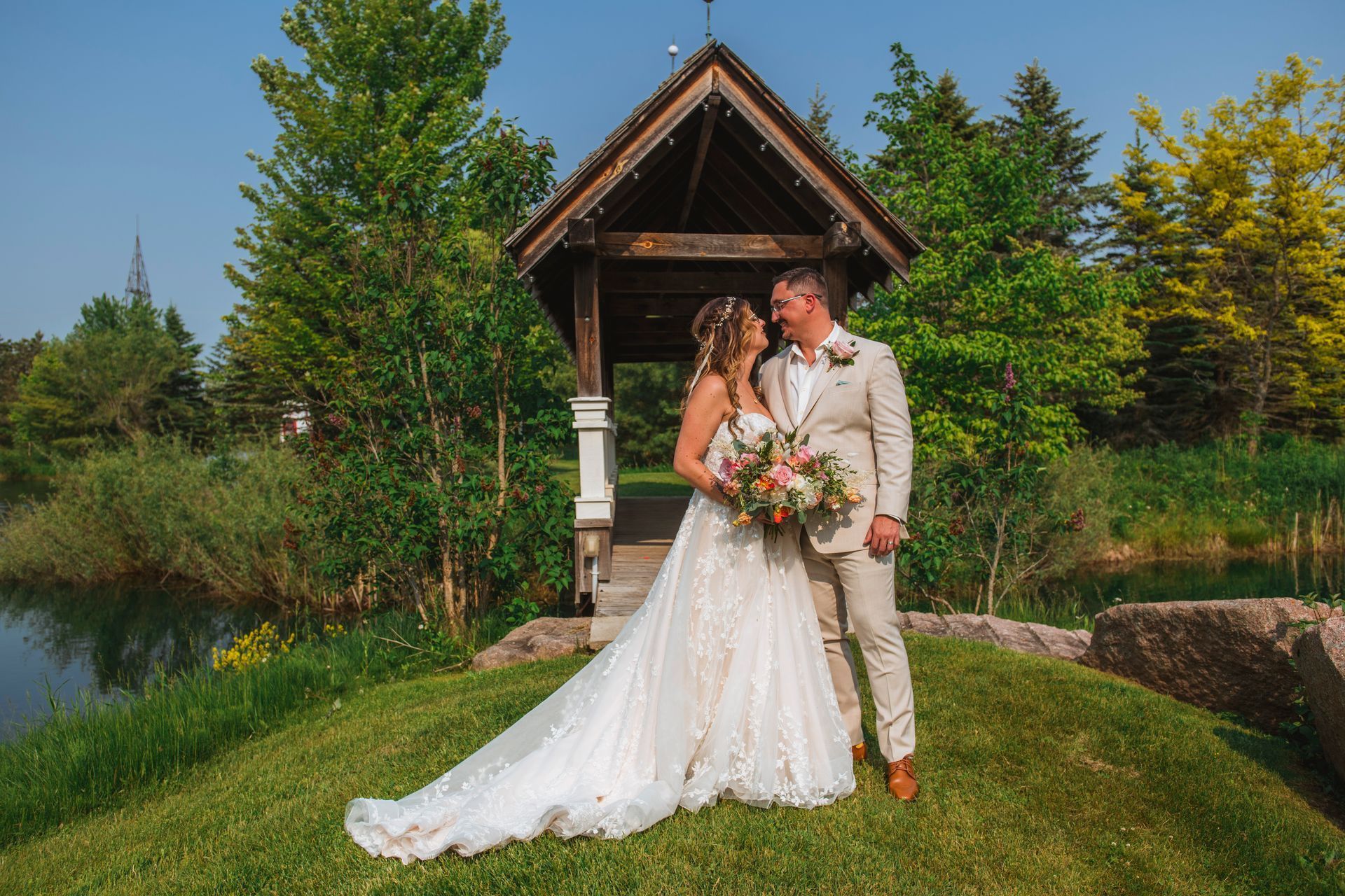 Bride and groom posing in wedding attire beside a small wooden chapel in a lush garden