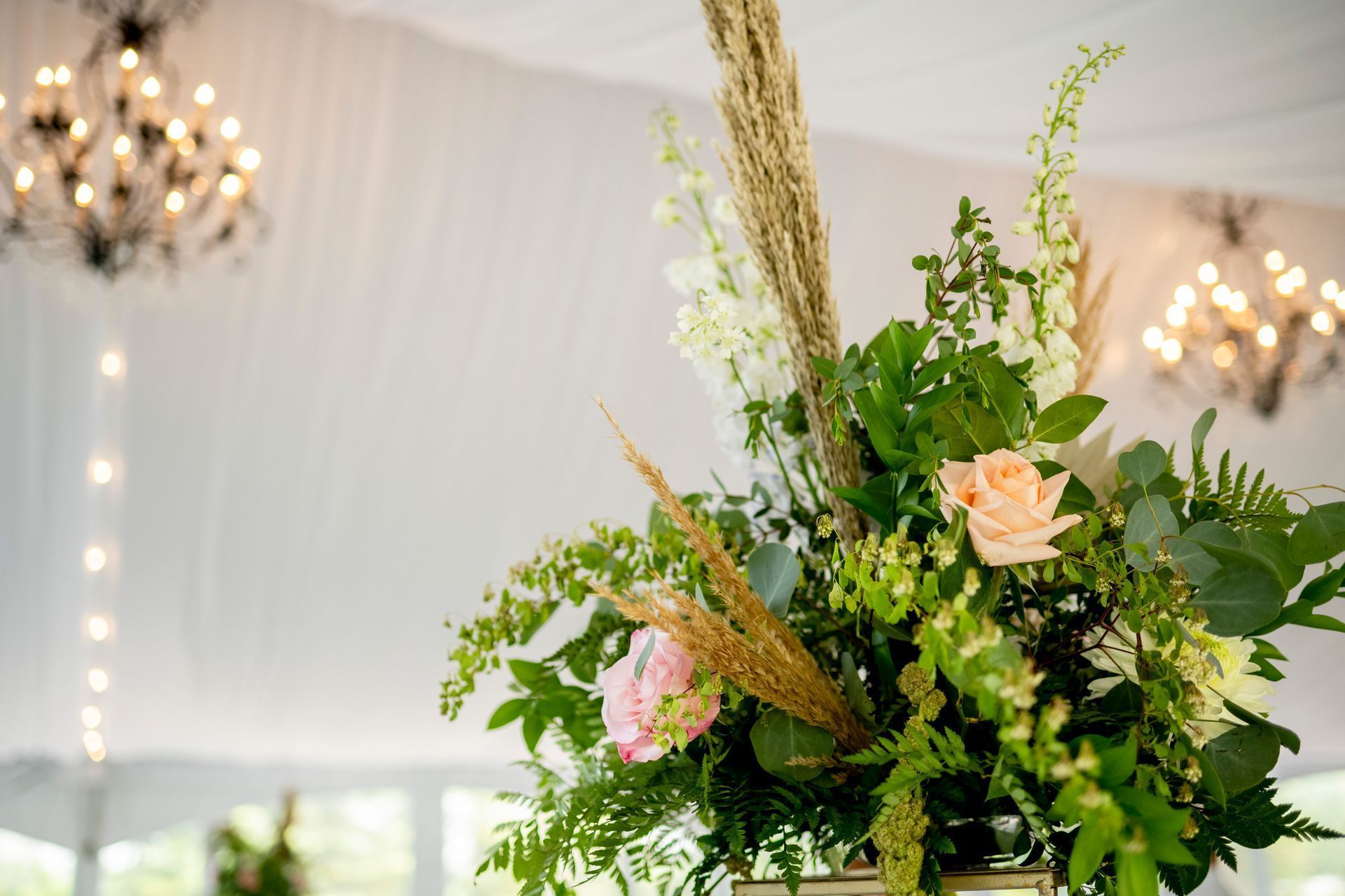 Floral arrangement with greenery and pale pink flowers in a bright, draped event tent