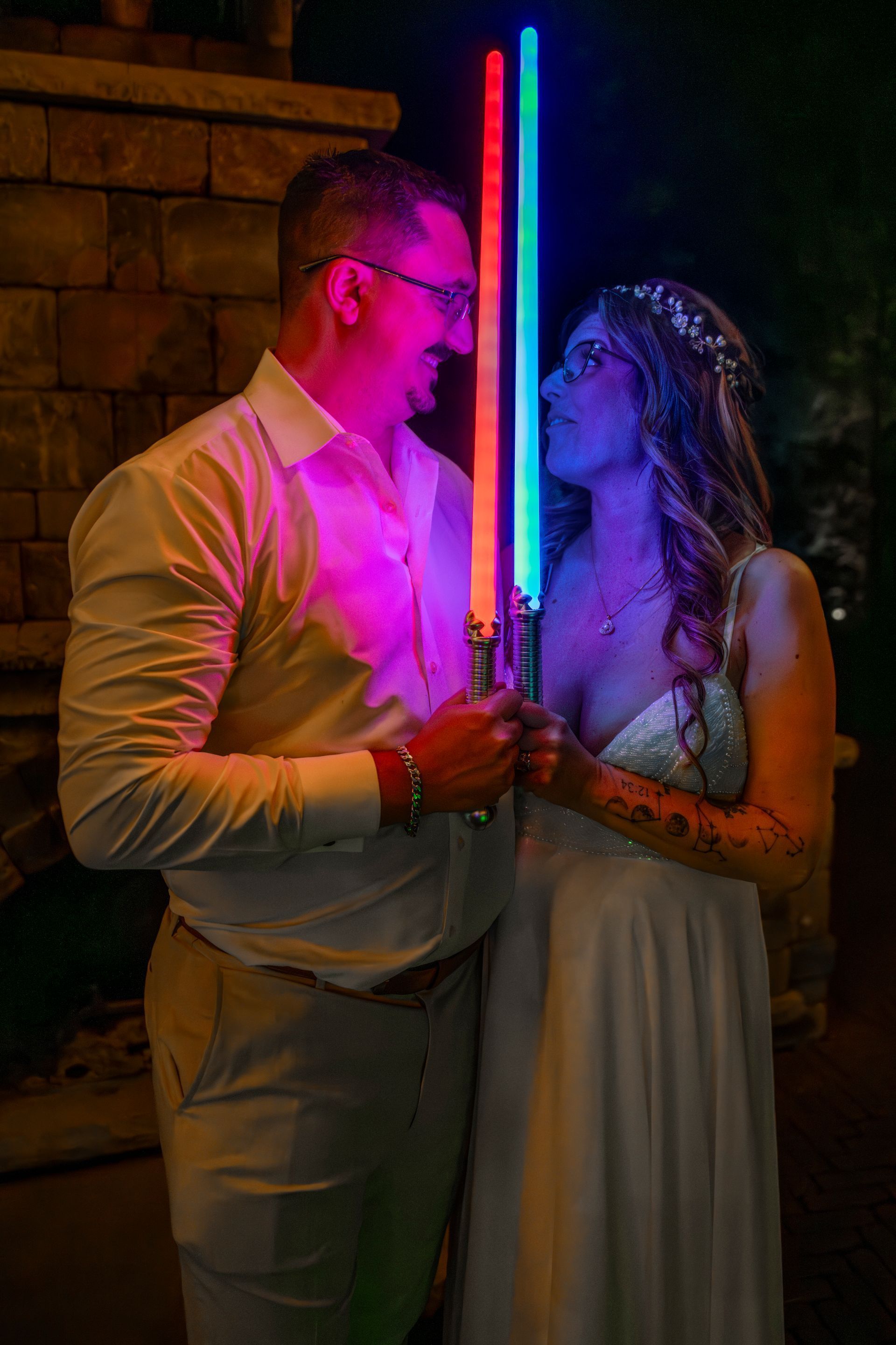 Couple holding glowing blue and pink lightsabers, facing each other at night outdoors