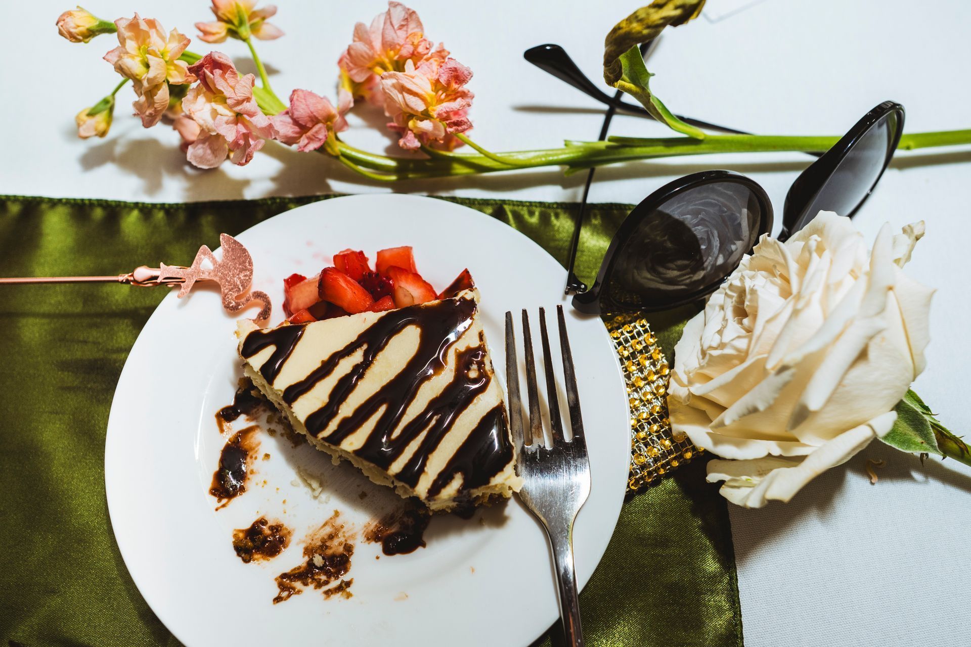 Dessert plate with chocolate-drizzled cake, strawberries, whipped cream, and sunglasses beside flowers on a green cloth