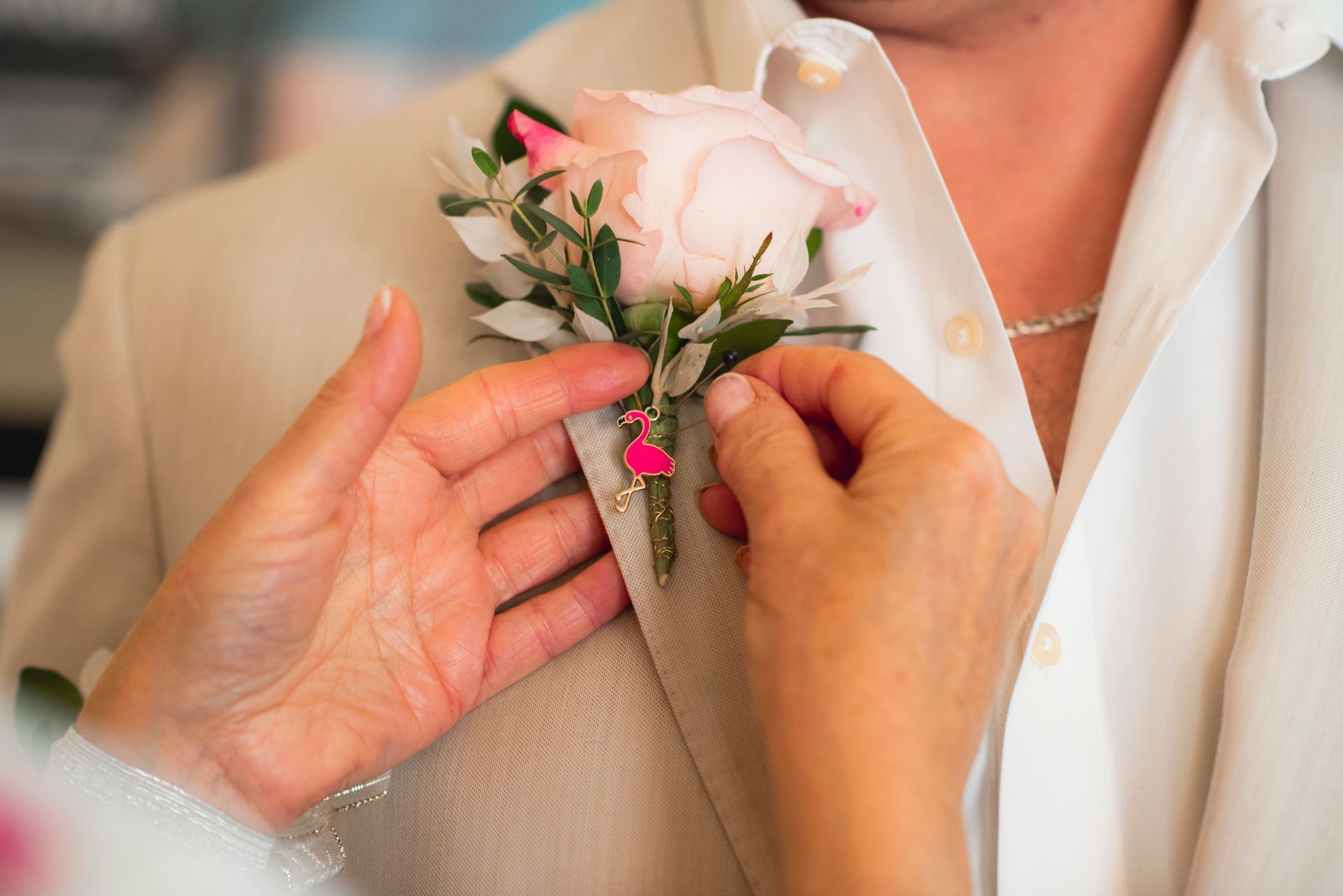 Hands pin a pink boutonniere onto a beige suit lapel over a white shirt.