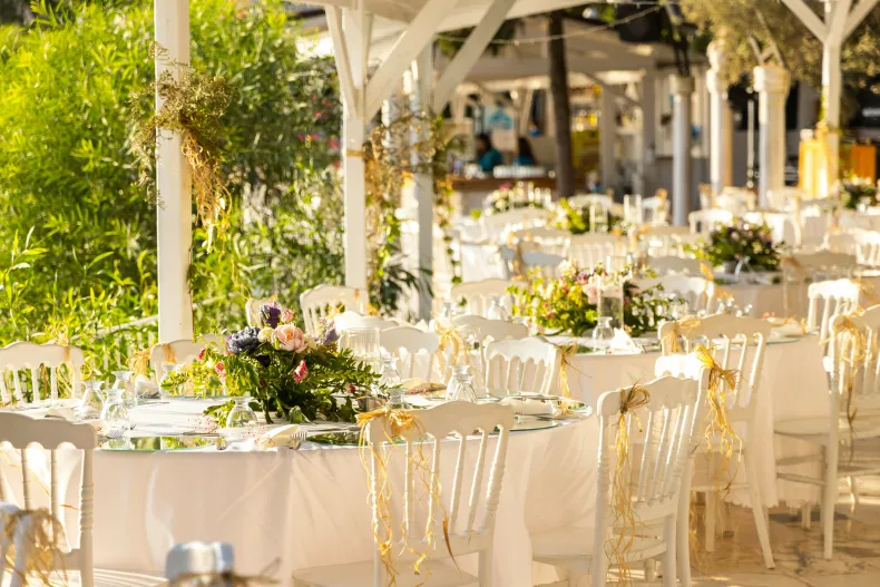 Sunlit outdoor wedding reception with white tables, gold chairs, and floral centerpieces under pergolas
