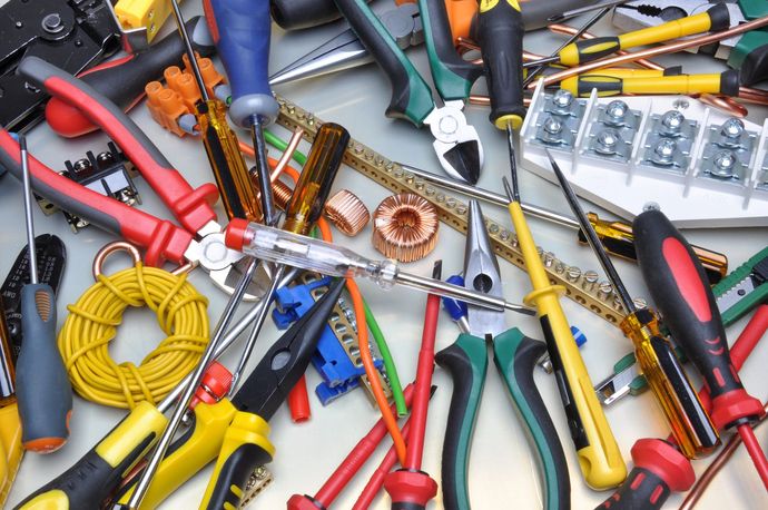 Su un banco da lavoro sono sparsi vari utensili manuali e materiale elettrico, tra cui cacciaviti, pinze, fili e connettori.