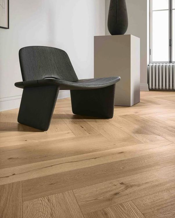 Poltrona moderna su pavimento in legno a spina di pesce accanto a un piedistallo e un vaso in una stanza luminosa.