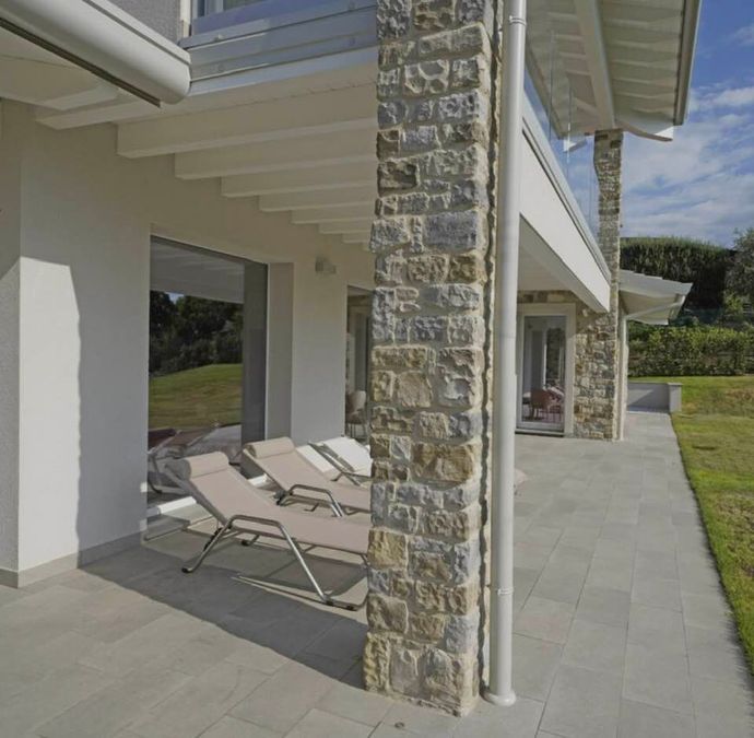 Patio coperto con sedie a sdraio accanto a una colonna in pietra e prato curato, sotto una moderna copertura.