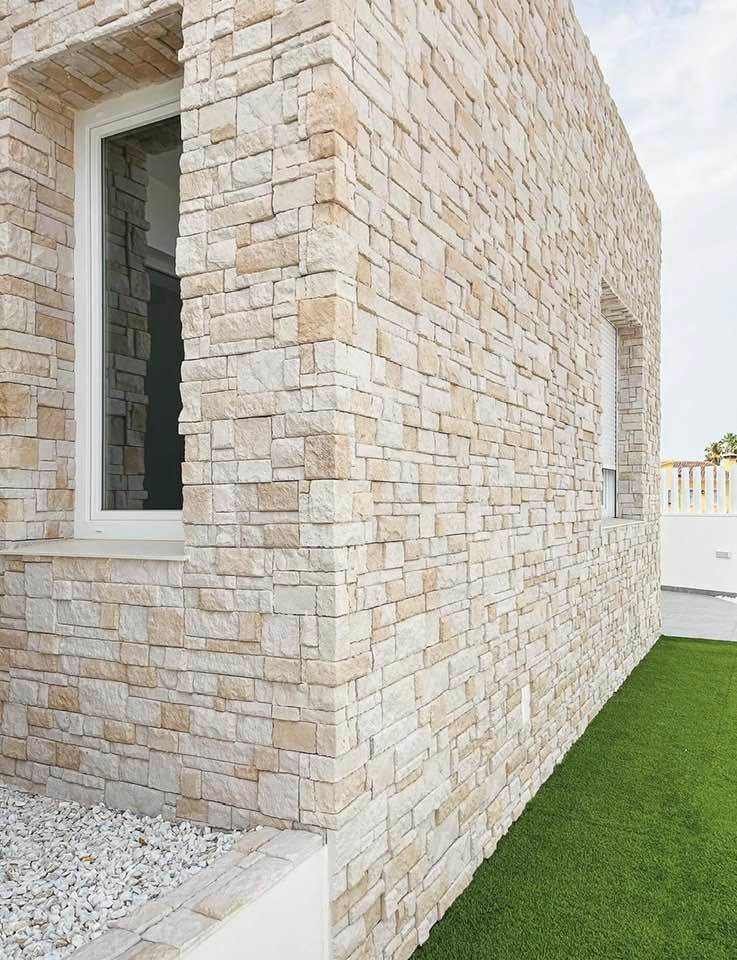 Muro esterno in pietra di una casa moderna con finestra e prato verde adiacente.