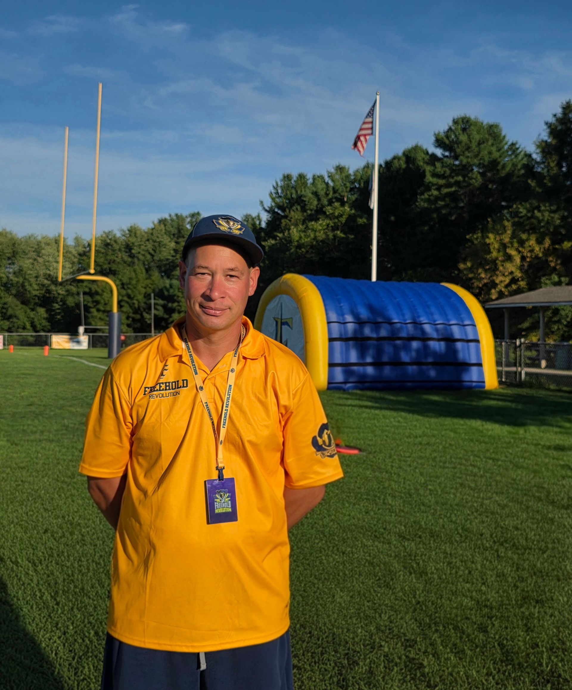 Football coach, arms crossed, on the field. Blue shirt, serious expression, players in background.