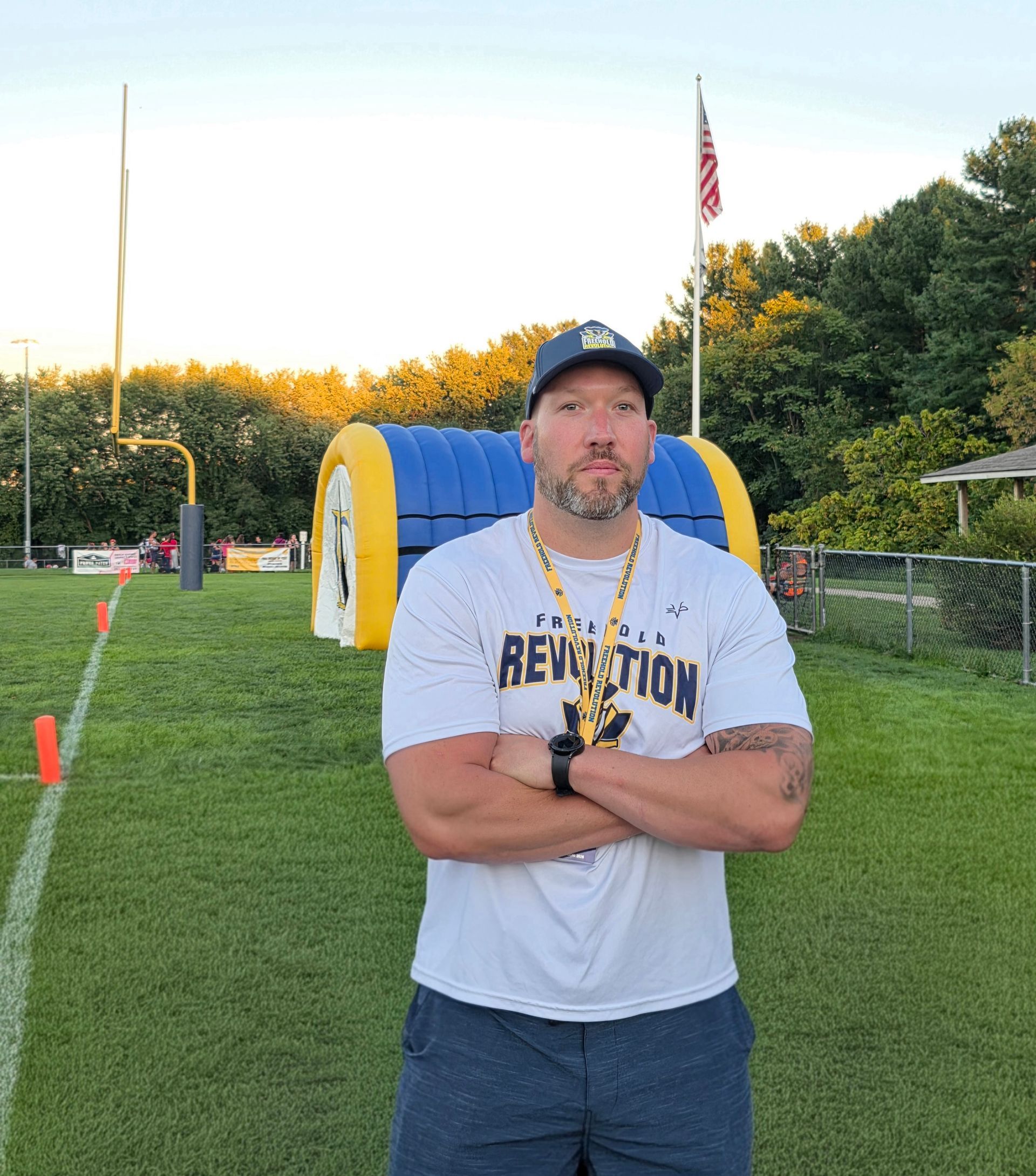 Man with crossed arms on a football field. He is wearing a blue polo shirt, khaki pants, and has a serious expression.