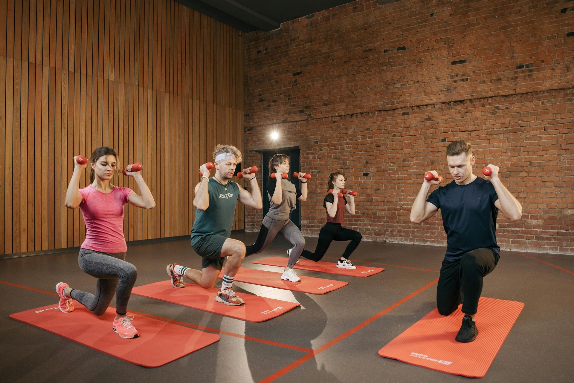 Five people doing lunges with dumbbells on red mats in a brick-walled gym.