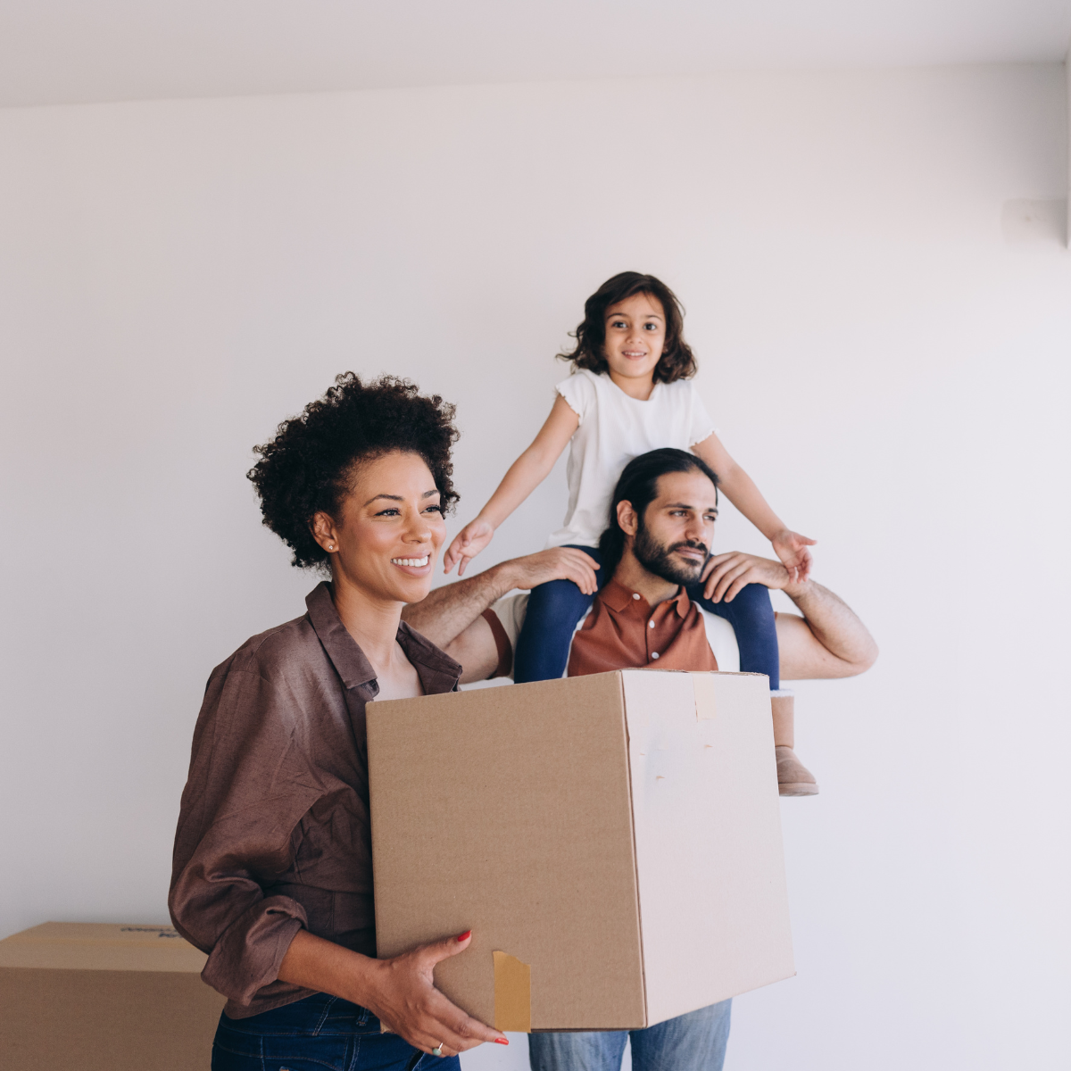 Family, smiling, carries cardboard box in new home; girl sits on dad's shoulders.