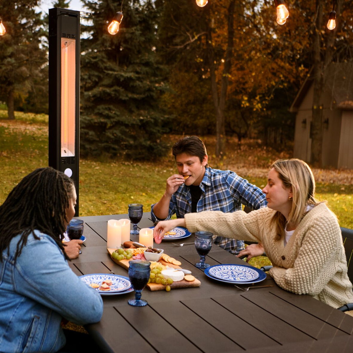 Three people dine at an outdoor table with a tall vertical heater, lit by string lights in a park setting.