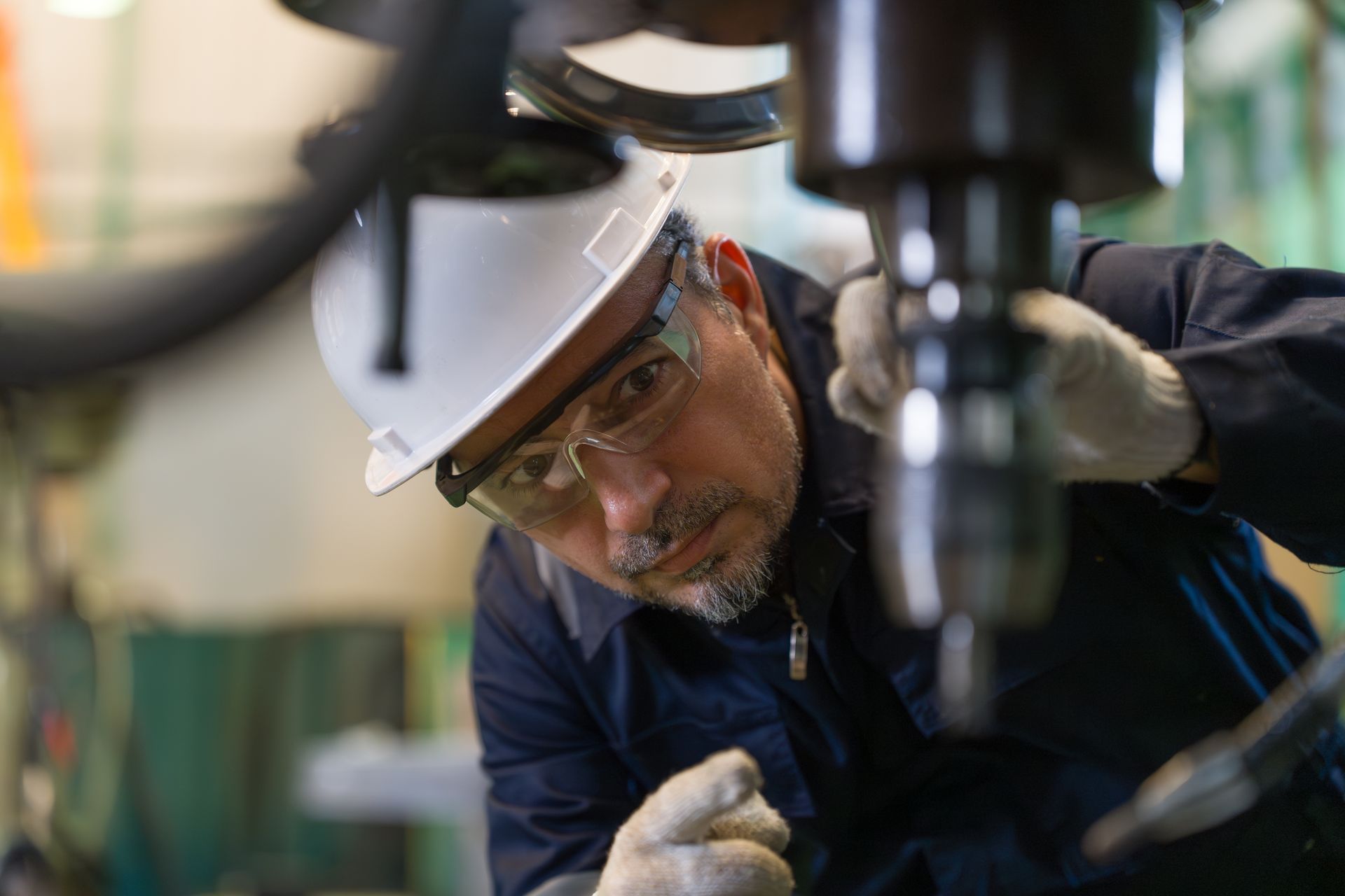 Worker in hard hat and safety glasses inspecting machinery in a factory, leaning in closely. Worker in hard hat and safety glasses inspecting machinery in a factory, leaning in closely.