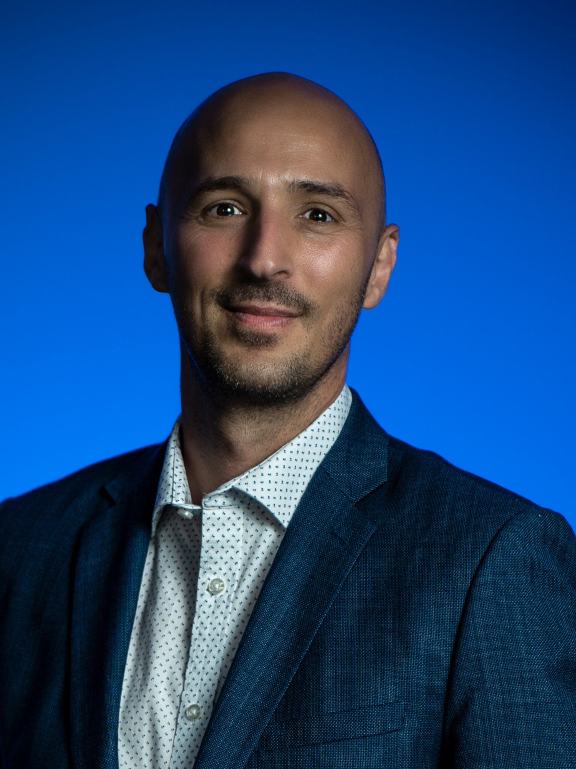 Portrait of a bald man in a blue suit and white shirt against a bright blue background