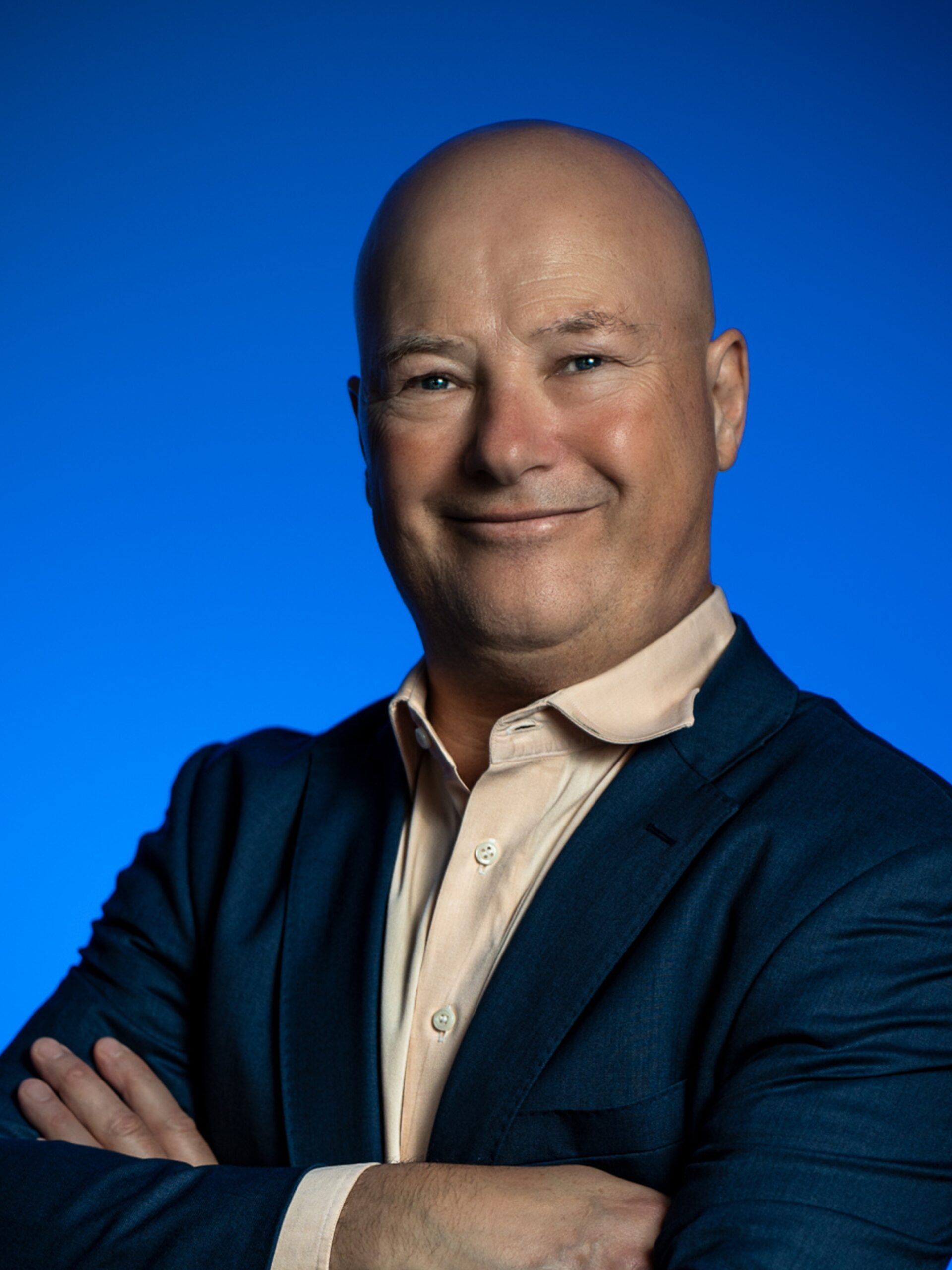 Smiling man in a beige shirt and blue sweater, arms crossed against a blue background