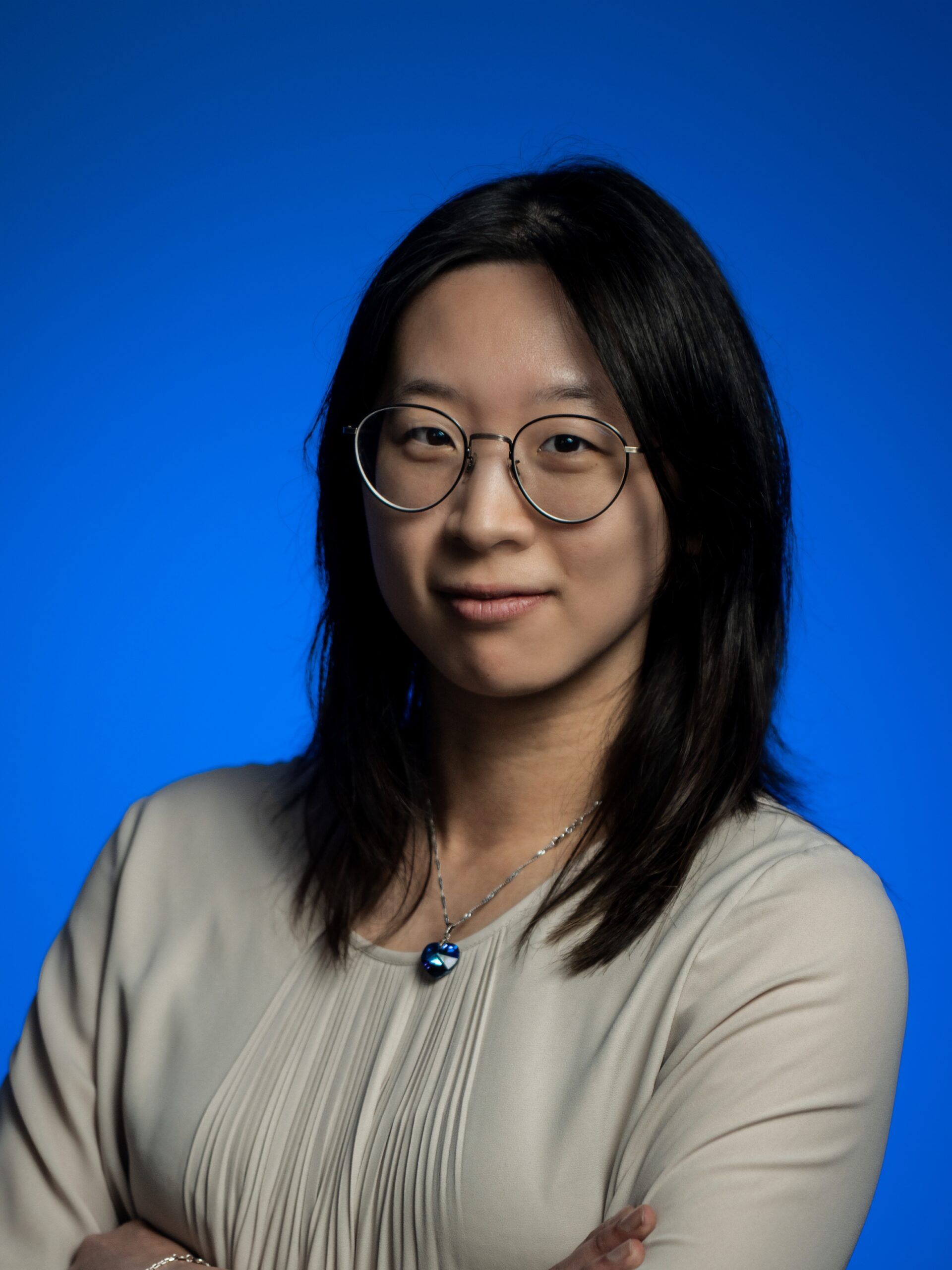 Portrait of a person with glasses against a blue studio background, wearing a beige top and a necklace