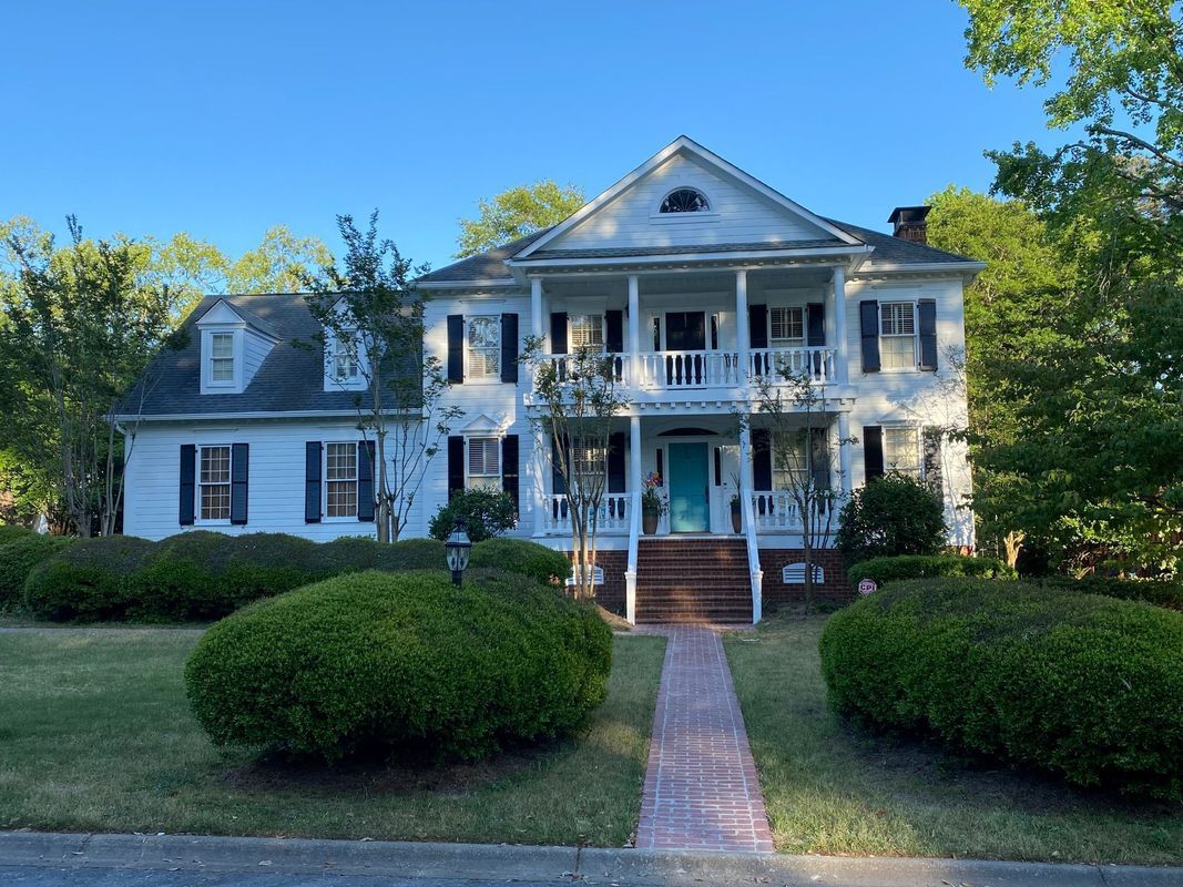 White two-story house with front porch and teal door, set on a green lawn with hedges.