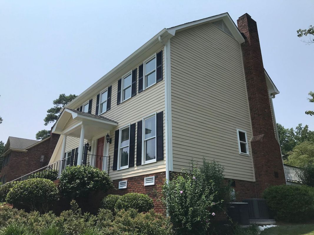 Two-story beige house with black shutters, brick chimney, and bushes in front. Blue sky background.