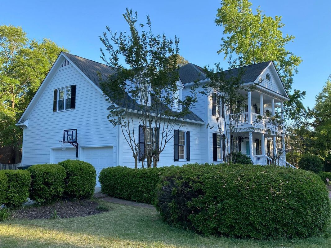White two-story house with black shutters, porch, and a basketball hoop. Green bushes and trees in the yard.