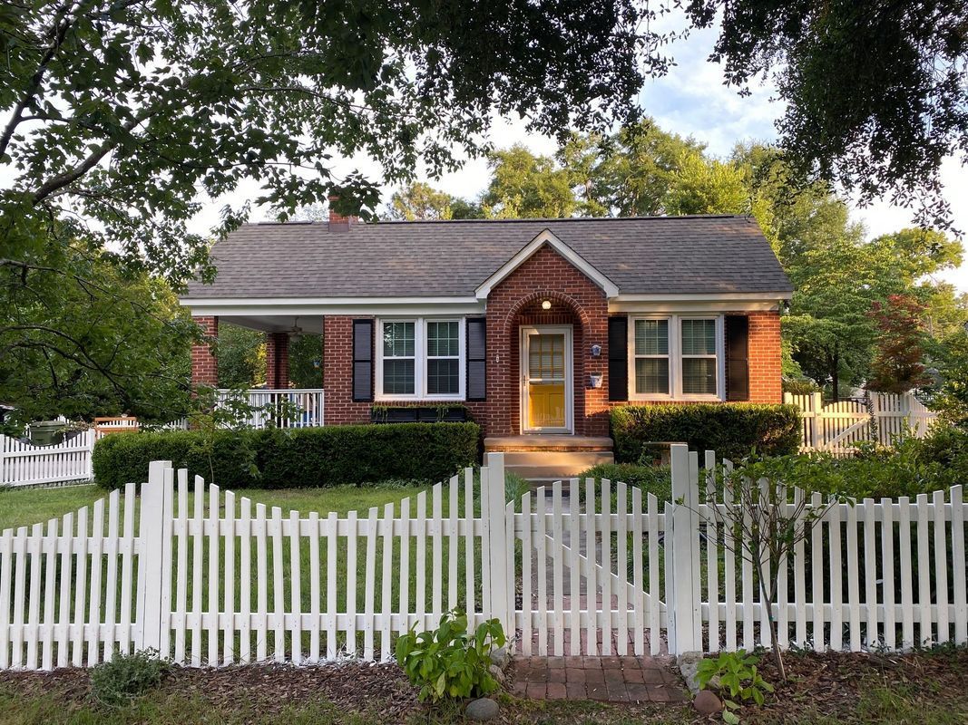 Cottage-style brick home with white picket fence, front porch, and black shutters, set among trees and green bushes.