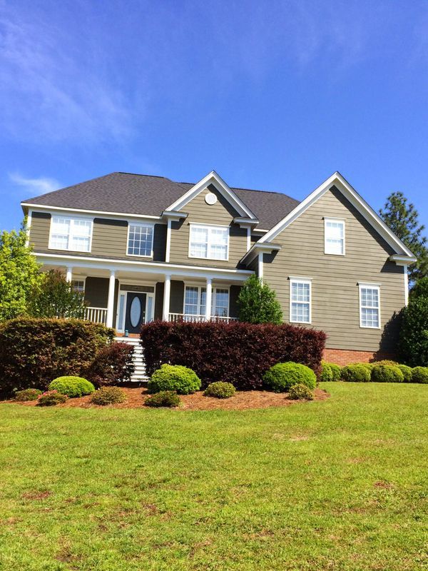 Green house with white trim, porch, and dark roof under a blue sky, surrounded by green grass and shrubs.