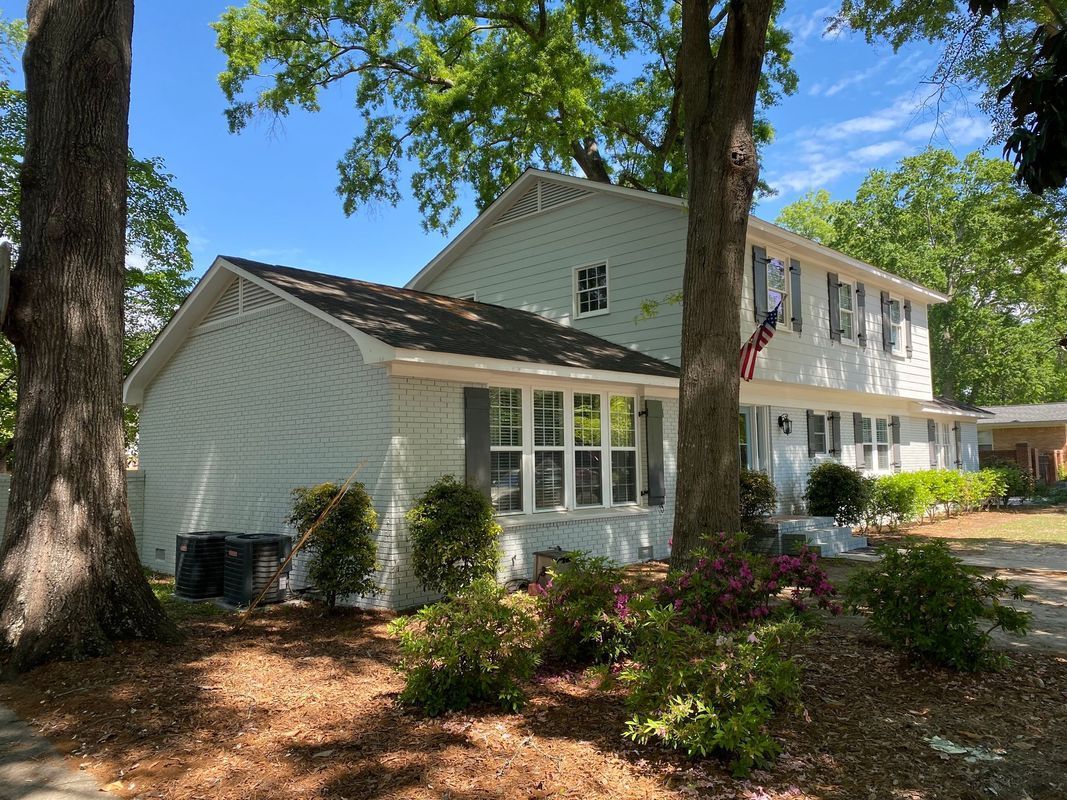 Two-story, light blue house with dark shutters, front porch, and small shrubs.