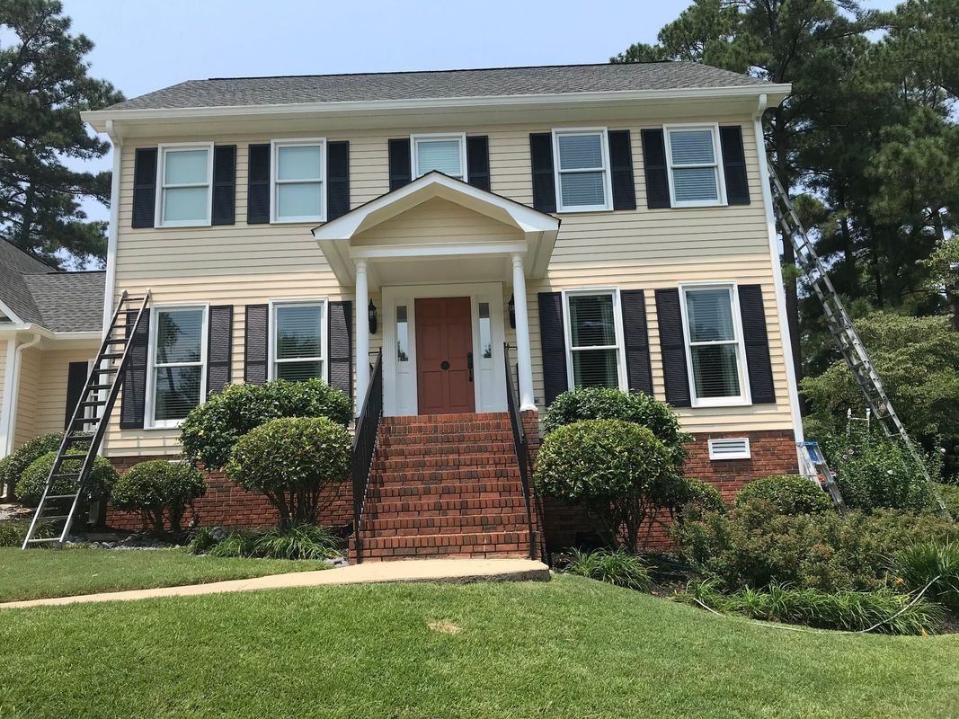 Two-story house with tan siding, black shutters, brick steps, and ladders leaning against the building.