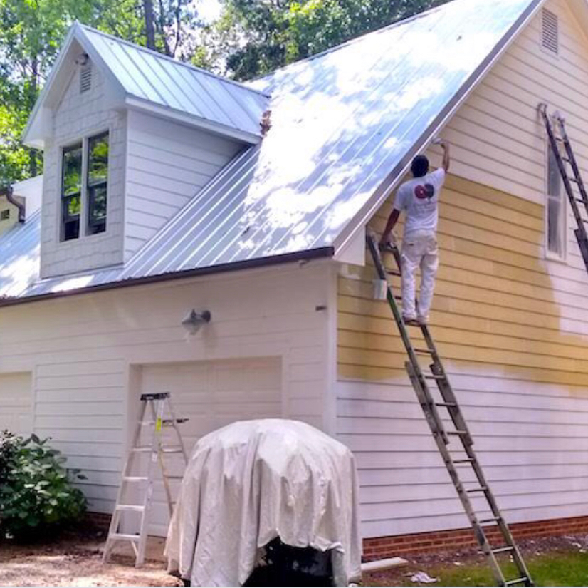 Person painting yellow siding on a white house, using a ladder. Metal roof and garage.