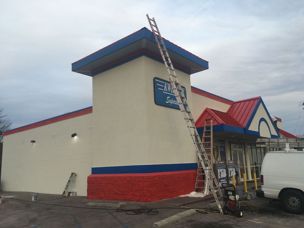 Building being painted, cream walls with red and blue trim, ladders propped against it.