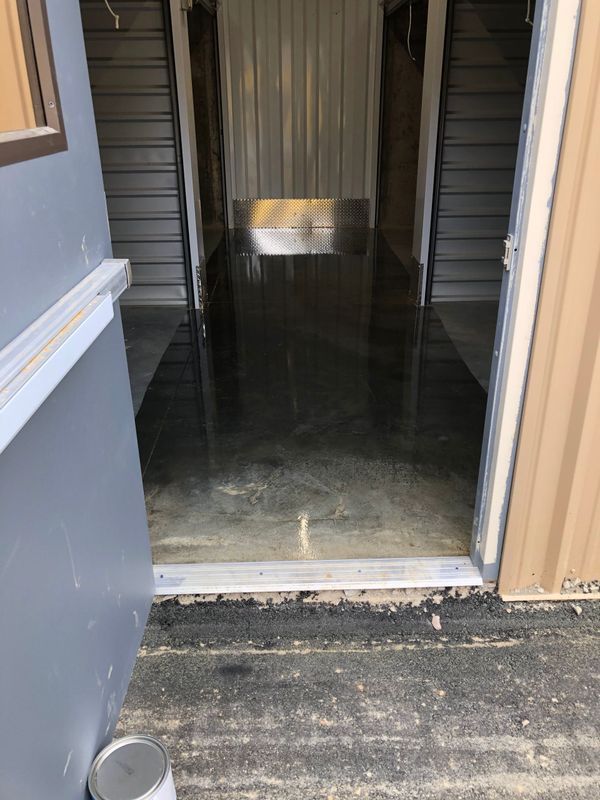 A storage unit with a reflective, wet concrete floor. The entrance has a grey door and a concrete threshold.